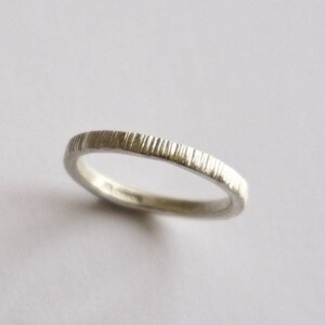 May include: A thin silver ring featuring a textured surface with vertical, parallel grooves etched along the entire outer band. The metal has a matte, metallic finish, and the ring is positioned at a slight angle to show its circular shape.