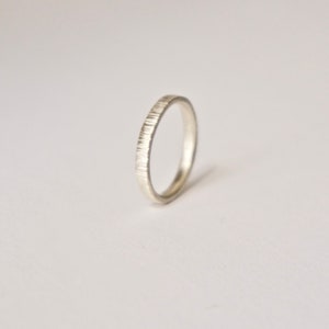 May include: A silver-colored ring with a thin band featuring a vertical, textured, etched pattern along the outer surface. The ring has a smooth, polished interior and a consistent, rounded shape, presented in a simple, clean, and neutral aesthetic.