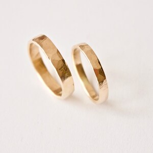 May include: Two gold rings with a hammered, textured finish. The rings are circular bands with a metallic yellow luster and uneven, faceted surfaces created by a hammering technique, showing a distinct contrast in width between the two pieces.