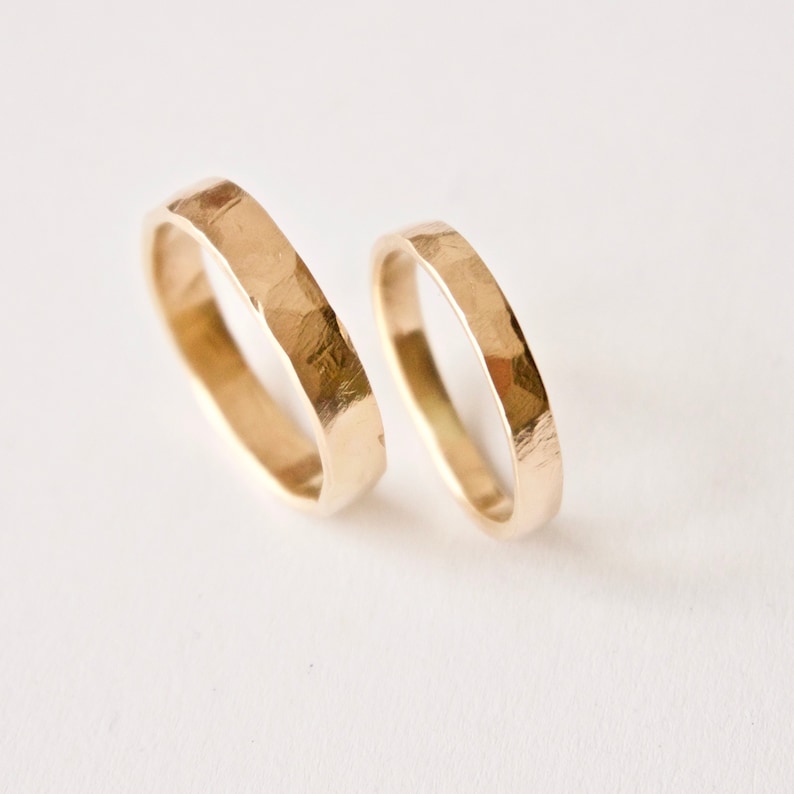 May include: Two gold rings with a hammered, textured finish. The rings are circular bands with a metallic yellow luster and uneven, faceted surfaces created by a hammering technique, showing a distinct contrast in width between the two pieces.