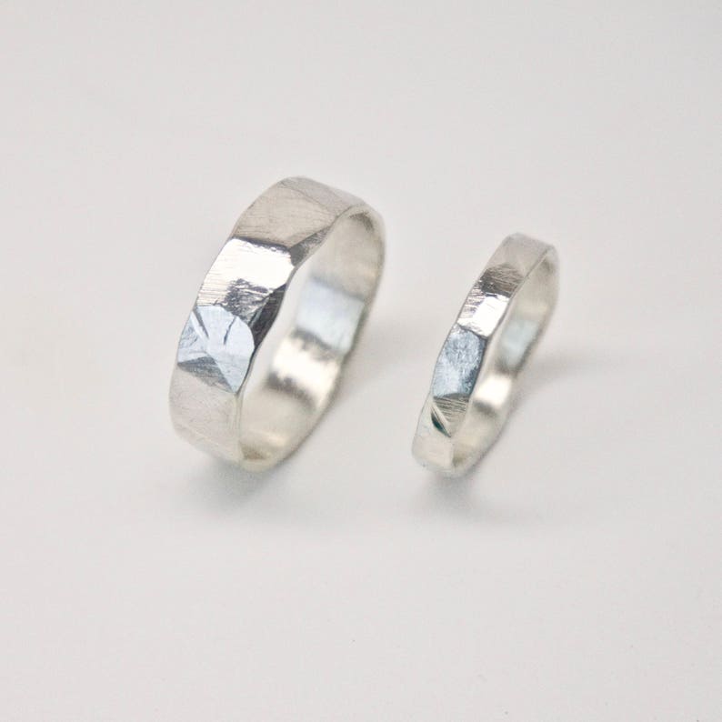 May include: Two silver rings with a faceted, textured design. The larger ring is wider than the smaller one. The rings are displayed on a white background, showcasing their metallic sheen and geometric texture. Ideal for couples.