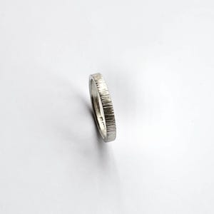 May include: A silver-colored metal ring with a textured, vertical line pattern on the outer band. The ring is positioned at an angle, revealing a smooth interior surface and a series of small, stamped hallmarks visible on the inner band.