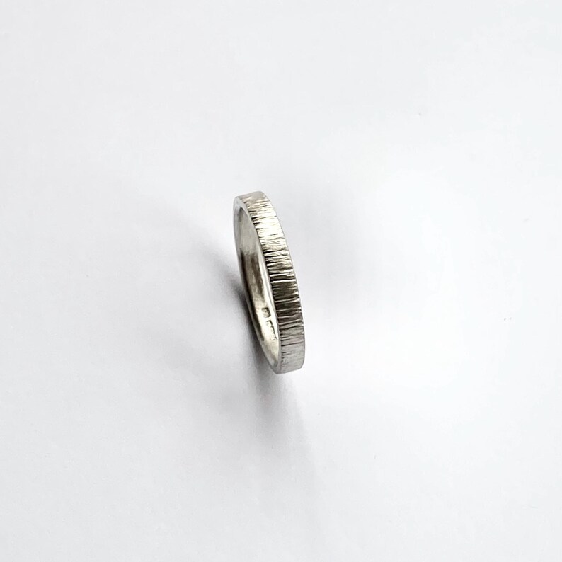 May include: A silver-colored metal ring with a textured, vertical line pattern on the outer band. The ring is positioned at an angle, revealing a smooth interior surface and a series of small, stamped hallmarks visible on the inner band.