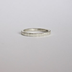 May include: A silver-colored ring with a thin band featuring a vertical, textured, grooved pattern along the outer surface. The metal has a matte, slightly brushed finish, creating a consistent appearance around the entire circumference of the band.