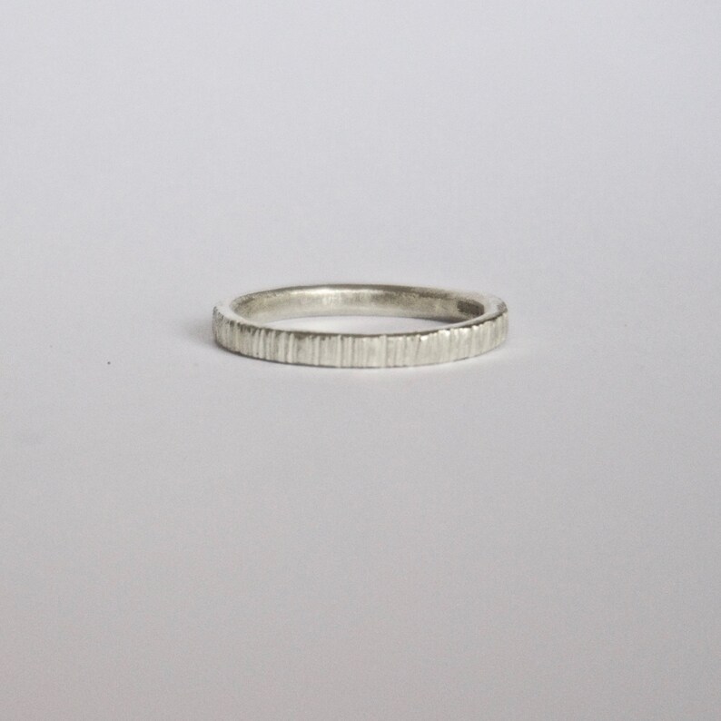 May include: A silver-colored ring with a thin band featuring a vertical, textured, grooved pattern along the outer surface. The metal has a matte, slightly brushed finish, creating a consistent appearance around the entire circumference of the band.
