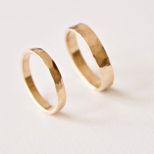 May include: Two gold rings with a hammered, textured finish. The rings feature a simple, circular band design with a reflective metallic surface. The smaller ring is positioned in the foreground, while the larger ring is placed slightly behind it.