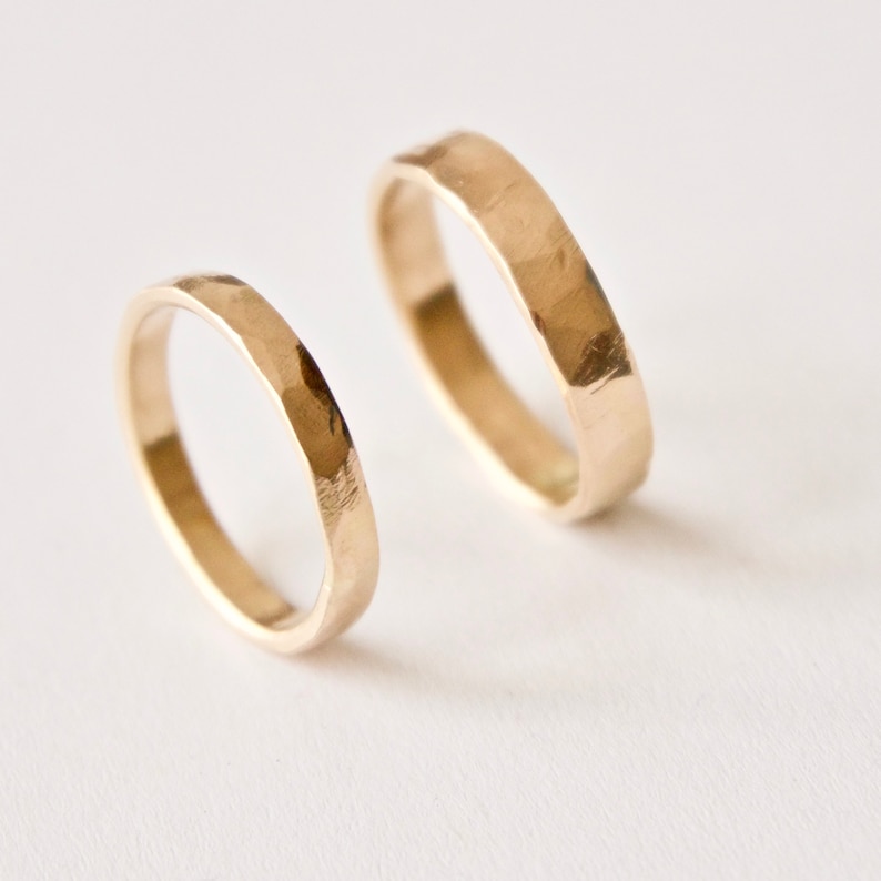 May include: Two gold rings with a hammered, textured finish. The rings feature a simple, circular band design with a reflective metallic surface. The smaller ring is positioned in the foreground, while the larger ring is placed slightly behind it.