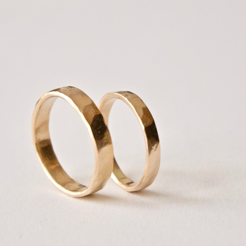 May include: Two gold-colored rings with a hammered, textured surface. The rings are circular with a polished finish that reflects light, showing irregular facets along the outer band. The rings are positioned upright, one slightly behind the other.