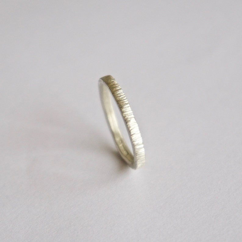 May include: A thin silver ring with a textured, vertical line pattern on the outer surface. The band has a metallic, light gray finish and a consistent width throughout its circumference, showcasing a detailed, etched appearance on the exterior.