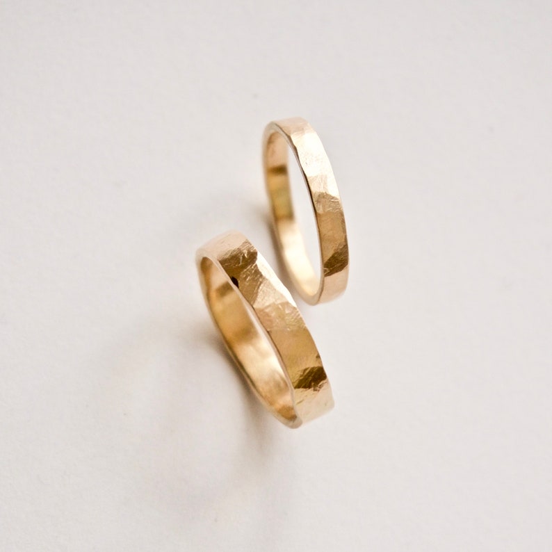 May include: Two gold rings with a distinct hammered texture on the outer surface. The bands have a polished finish that reflects light, creating a bright appearance. The rings are positioned at slight angles to each other, showcasing the metal work.