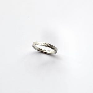 May include: A silver-colored metal ring with a textured outer surface featuring vertical etched lines. The band has a smooth interior finish and a consistent width throughout its circumference, presenting a metallic sheen against a plain background.