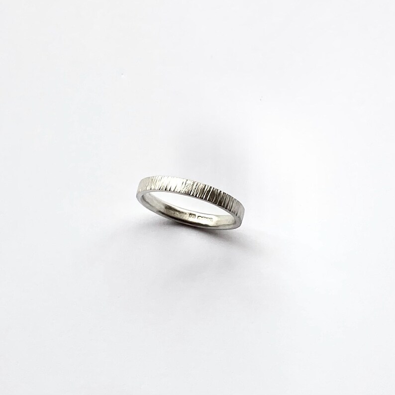May include: A silver-colored metal ring with a textured outer surface featuring vertical etched lines. The band has a smooth interior finish and a consistent width throughout its circumference, presenting a metallic sheen against a plain background.