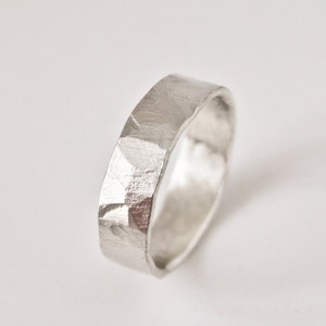 May include: A silver-colored ring with a wide band and a textured, hammered finish. The surface of the metal features irregular, faceted indentations that catch the light, creating a matte and reflective appearance across the entire outer circumference.