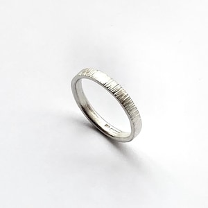 May include: A silver-colored ring with a textured, vertical-line finish on the outer band. The interior of the band features small, stamped hallmarks visible near the bottom edge, indicating metal purity and origin, set against a plain white background.