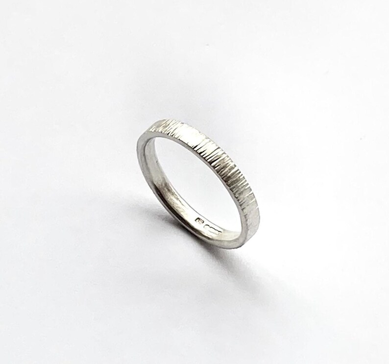 May include: A silver-colored ring with a textured, vertical-line finish on the outer band. The interior of the band features small, stamped hallmarks visible near the bottom edge, indicating metal purity and origin, set against a plain white background.