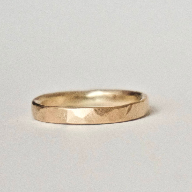 May include: A gold ring with a hammered finish. The ring features a flat band and a gently curved inner surface. The textured surface of the ring catches the light, producing highlights and shadows. A simple, elegant piece of jewellery.