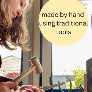 May include: A light-colored wooden mallet and a metal ring mandrel are used to shape a metal ring. A circular yellow graphic overlay contains the black text: made by hand using traditional tools. The scene shows metalworking equipment in use.