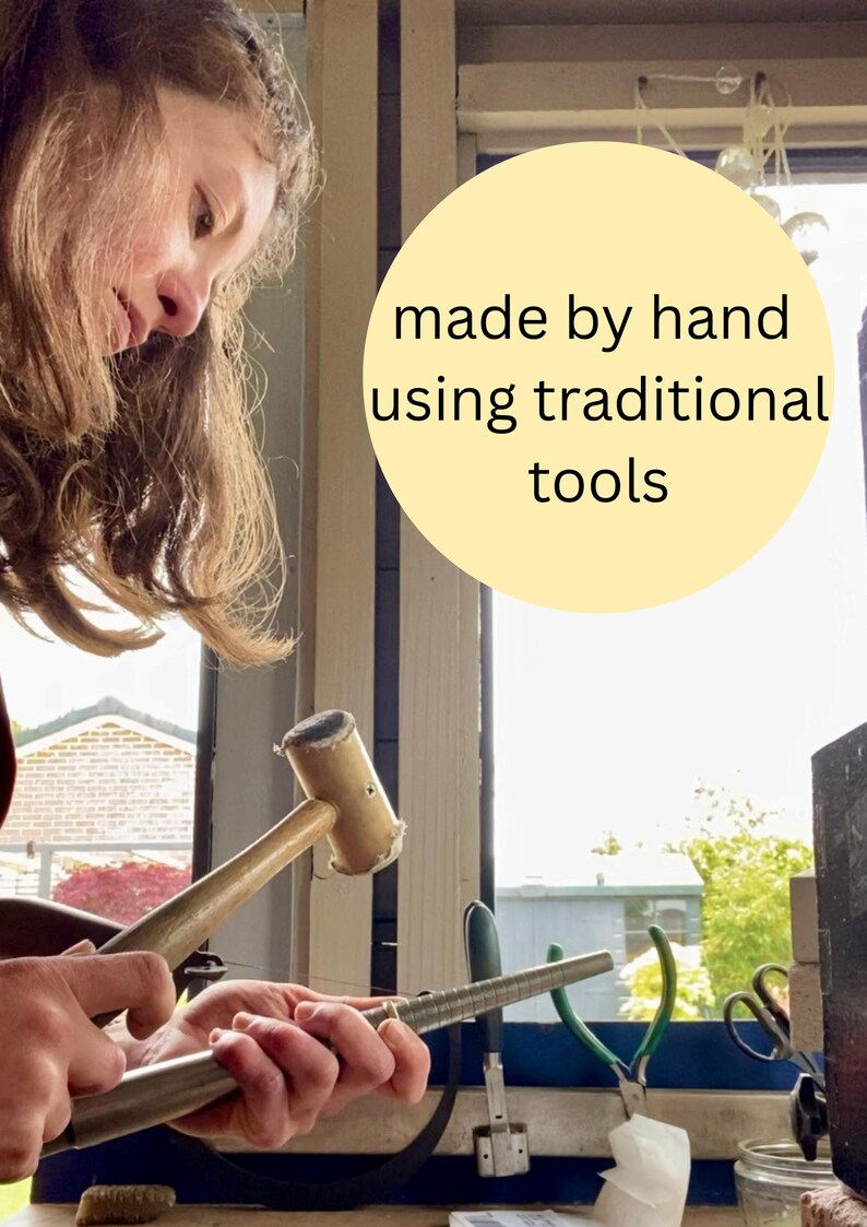 May include: A light-colored wooden mallet and a metal ring mandrel are used to shape a metal ring. A circular yellow graphic overlay contains the black text: made by hand using traditional tools. The scene shows metalworking equipment in use.