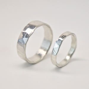 May include: Two silver rings with a textured, faceted surface. The larger ring is thicker than the smaller one. The rings are set against a plain white background, highlighting their metallic sheen. These rings are likely for a wedding or engagement.