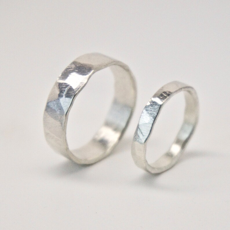 May include: Two silver rings with a textured, faceted surface. The larger ring is thicker than the smaller one. The rings are set against a plain white background, highlighting their metallic sheen. These rings are likely for a wedding or engagement.