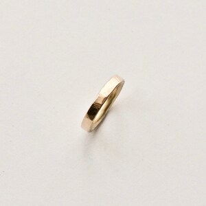 May include: A gold-coloured ring with a hammered texture. The ring is a simple band design, with a slightly irregular surface. The ring is set against a white background, highlighting its metallic sheen. The ring appears to be approximately 0.25 cm wide.