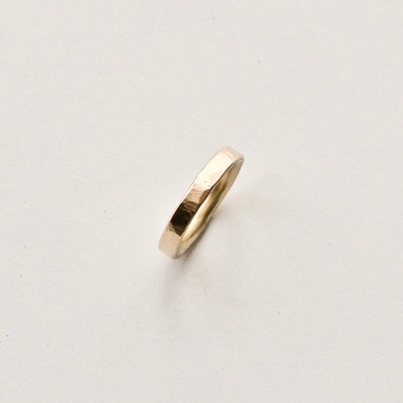 May include: A gold-coloured ring with a hammered texture. The ring is a simple band design, with a slightly irregular surface. The ring is set against a white background, highlighting its metallic sheen. The ring appears to be approximately 0.25 cm wide.