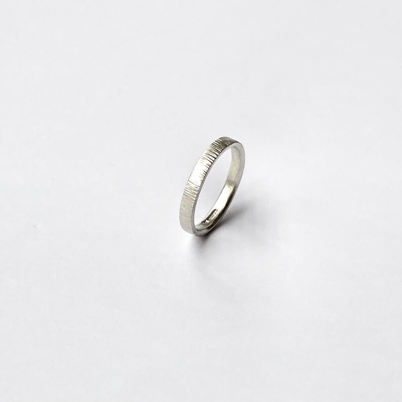 May include: A silver-colored ring with a textured, vertical line pattern on the outer surface. The band has a smooth, polished interior and a consistent width throughout, showcasing a metallic finish that reflects light across its surface.