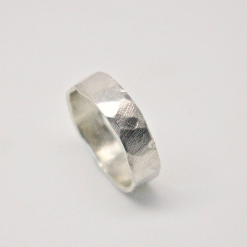 May include: A silver ring with a hammered texture. The ring has a flat band and a faceted surface, reflecting light. The ring is shown on a white background.