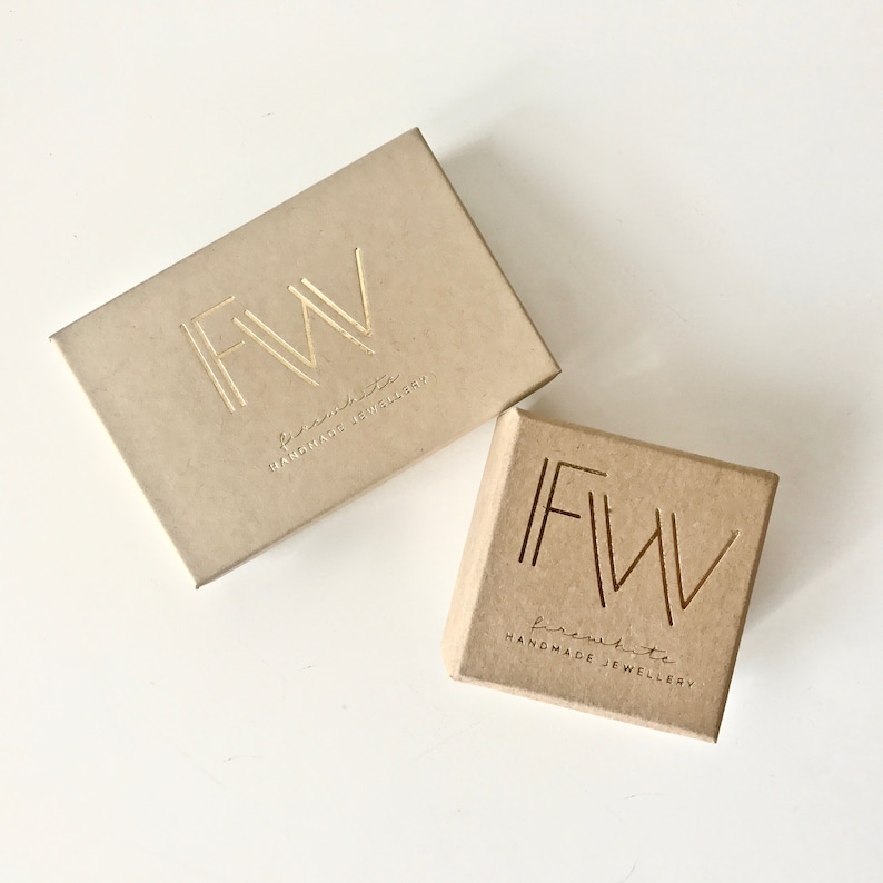May include: Two rectangular cardboard jewelry boxes in a light tan color. Each box features the gold-stamped logo FWV with the text firewhite HANDMADE JEWELLERY printed underneath. The boxes are positioned at an angle on a plain white surface.