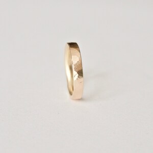 May include: A gold ring with a hammered finish. The ring has a simple band design, with a slightly rounded inside and a flat outside. The ring is set against a white background.