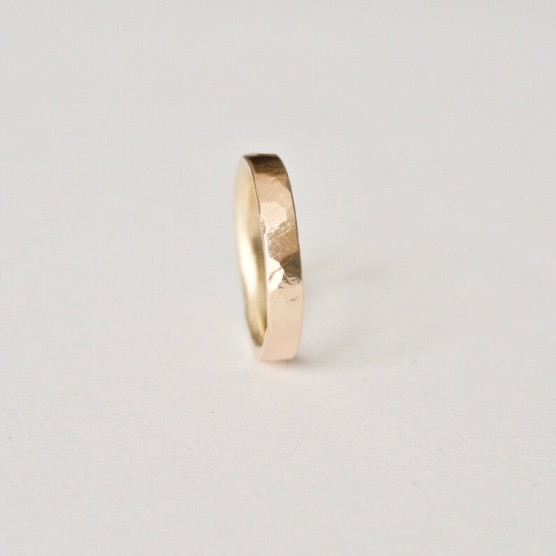 May include: A gold ring with a hammered finish. The ring has a simple band design, with a slightly rounded inside and a flat outside. The ring is set against a white background.