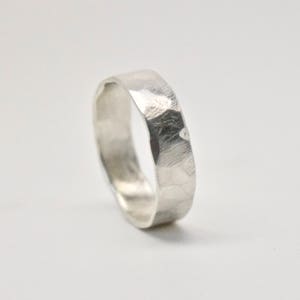 May include: A silver ring with a hammered texture. The ring has a flat band and a slightly irregular surface, reflecting light. The ring is set against a white background, highlighting its metallic sheen. A simple, elegant piece of jewelry.