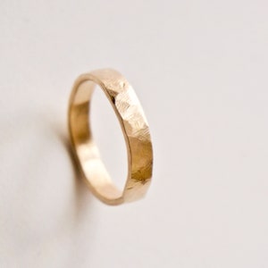 May include: A gold ring with a textured, hammered finish on the outer surface. The band has a rounded interior and a slightly irregular, faceted exterior appearance, reflecting light across its metallic surface in a warm, golden tone.