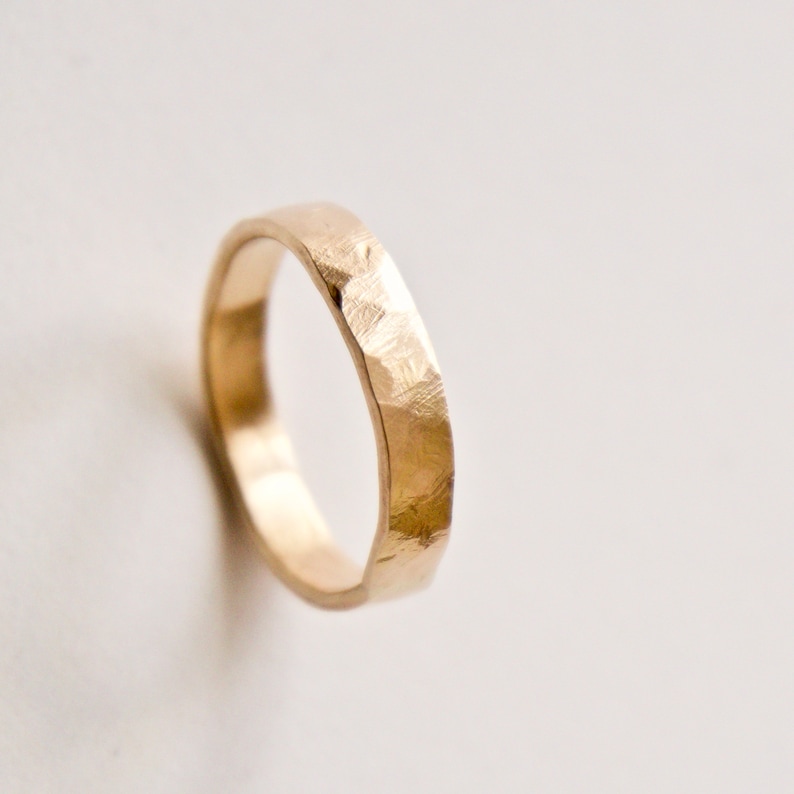 May include: A gold ring with a textured, hammered finish on the outer surface. The band has a rounded interior and a slightly irregular, faceted exterior appearance, reflecting light across its metallic surface in a warm, golden tone.