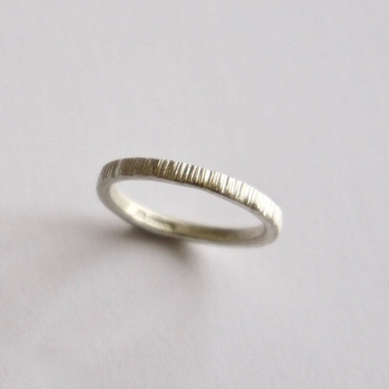 May include: A thin silver ring featuring a textured surface with vertical, parallel grooves etched along the entire outer band. The metal has a matte, metallic finish, and the ring is positioned at a slight angle to show its circular shape.
