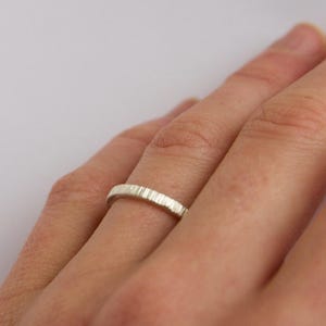 May include: A silver-colored ring with a textured, vertical-lined surface pattern. The band has a consistent width and a matte finish, featuring a series of fine, parallel grooves etched around the entire circumference of the metal ring.