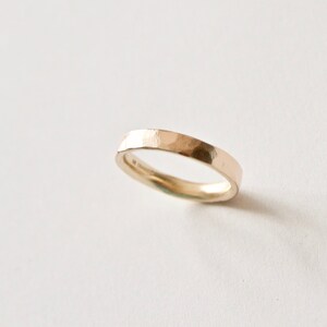 May include: A gold ring with a hammered texture. The band is flat and has a slightly irregular shape. The ring is set against a white background, highlighting its metallic sheen. A minimalist piece of jewellery.