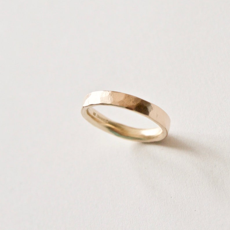 May include: A gold ring with a hammered texture. The band is flat and has a slightly irregular shape. The ring is set against a white background, highlighting its metallic sheen. A minimalist piece of jewellery.