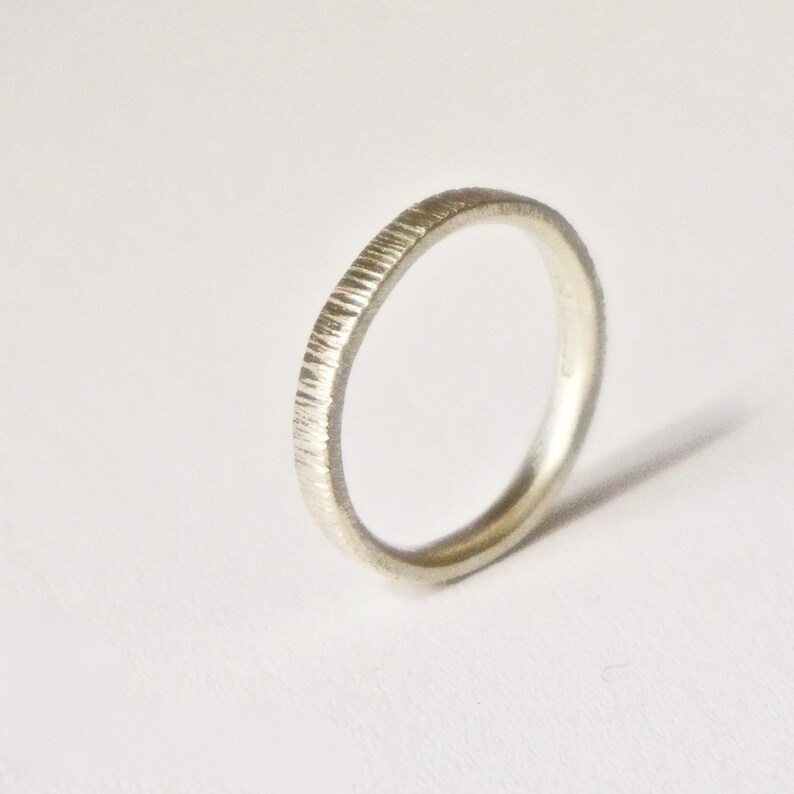 May include: A silver-toned ring with a textured, vertical-lined finish on the outer band. The ring has a slim, circular profile and a metallic sheen, showcasing a consistent pattern of fine, etched grooves around the entire circumference.