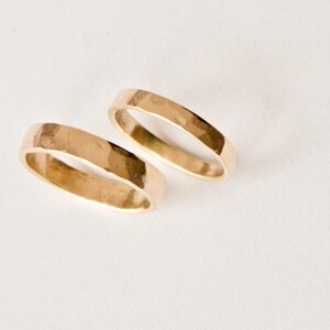 May include: Two gold-colored rings with a hammered, textured surface. Each ring features a flat, wide band design with a polished finish that reflects light, showcasing a consistent metallic yellow hue across the entire exterior surface.