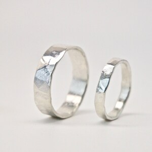 May include: Two silver rings with a textured, faceted design. The larger ring has a wider band, while the smaller ring has a thinner band. Both rings are set against a white background, highlighting their metallic sheen. Ideal for wedding or engagement.