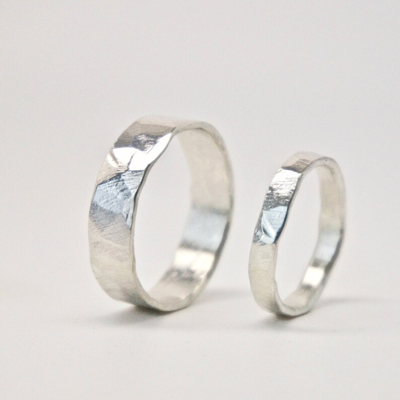 May include: Two silver rings with a textured, faceted design. The larger ring has a wider band, while the smaller ring has a thinner band. Both rings are set against a white background, highlighting their metallic sheen. Ideal for wedding or engagement.