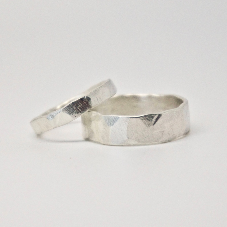 May include: Two silver rings with a hammered texture. One ring is thin, and the other is wider. The rings are set against a white background, highlighting their metallic sheen and unique surface detail. Ideal for weddings or everyday wear.