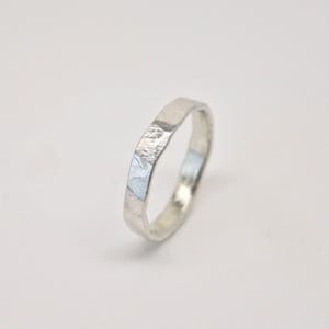 May include: A silver ring with a textured, faceted design. The ring has a band with a slightly irregular shape and a matte finish. The ring is shown against a white background, highlighting its metallic sheen.