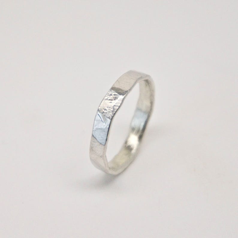May include: A silver ring with a textured, faceted design. The ring has a band with a slightly irregular shape and a matte finish. The ring is shown against a white background, highlighting its metallic sheen.