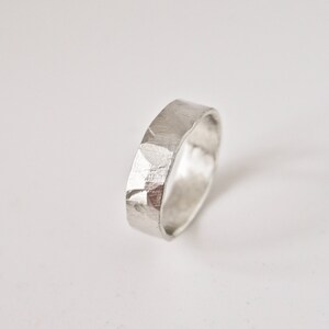 May include: A silver ring with a textured, faceted surface. The ring has a flat band design and a slightly hammered appearance. The ring is shown on a white background, highlighting its metallic sheen and simple elegance.