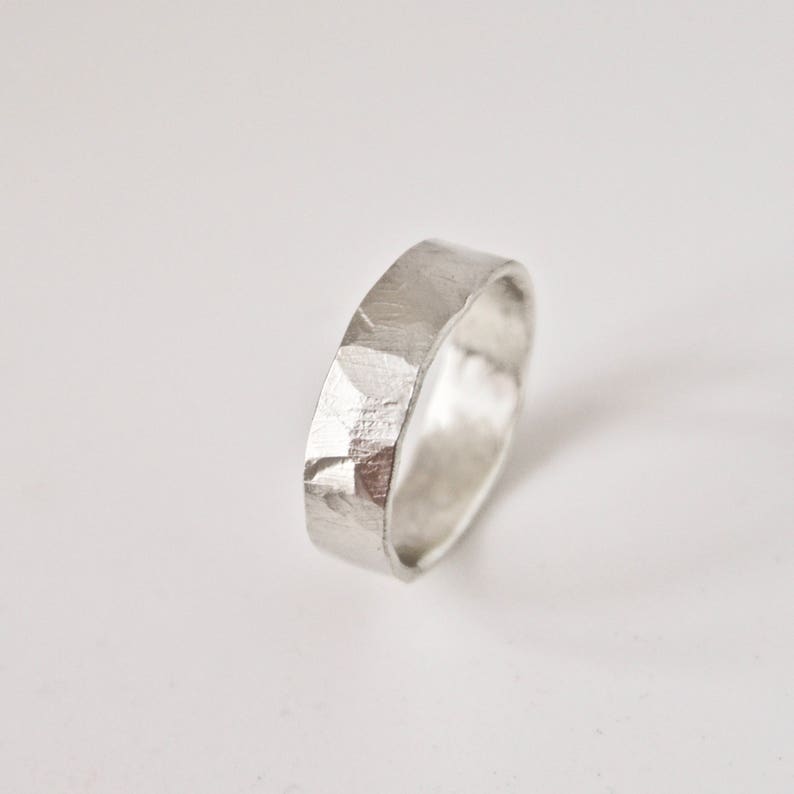 May include: A silver ring with a textured, faceted surface. The ring has a flat band design and a slightly hammered appearance. The ring is shown on a white background, highlighting its metallic sheen and simple elegance.