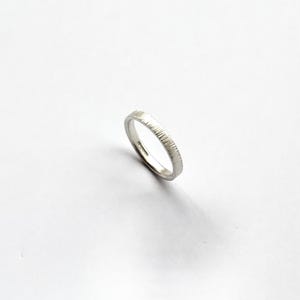 May include: A silver-colored ring with a textured outer surface featuring fine, vertical etched lines. The band has a smooth, polished interior and a consistent width throughout its circular shape, presented against a plain, neutral background.