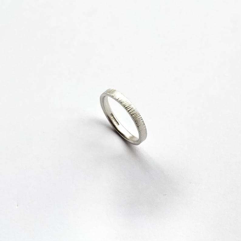May include: A silver-colored ring with a textured outer surface featuring fine, vertical etched lines. The band has a smooth, polished interior and a consistent width throughout its circular shape, presented against a plain, neutral background.