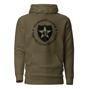 Puede incluir: Una sudadera con capucha verde con un gráfico en blanco y negro del emblema de la 2.ª División de Infantería. El emblema presenta una estrella, un escudo y una cabeza de indio americano. El texto alrededor del emblema dice "2nd Infantry Division Camp Red Cloud South Korea".